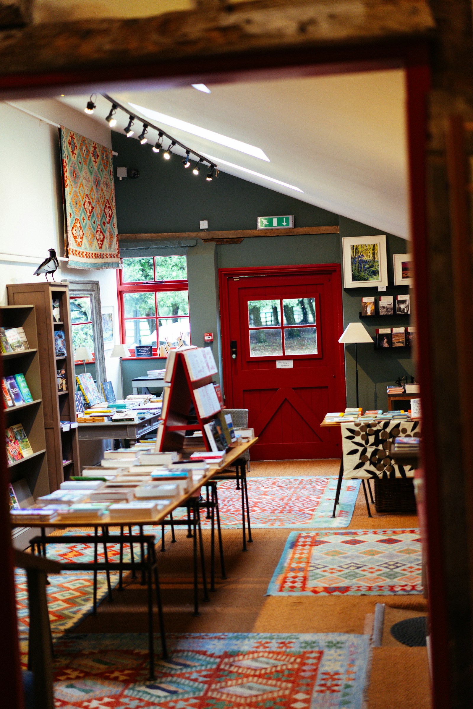 Warm, cosy independent bookshop interior with book-laden tables, patterned rugs and a red door